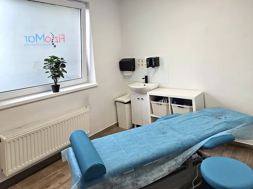 Sala terapii w gabinecie FizjoMar przygotowana na pacjenta: elektryczny stół rehabilitacyjny z podkładem i wałkiem, taboret, umywalka i okno z logo FizjoMar