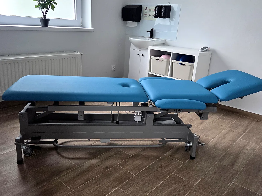 Sala terapii w gabinecie FizjoMar: elektryczny stół rehabilitacyjny, umywalka, regał i okno z logo FizjoMar